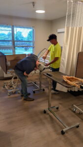 Two people work together assembling or repairing a piece of medical equipment in a room with large windows and medical curtains. Tools and parts are on a rolling table nearby.