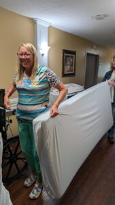 A smiling woman stands in a hallway, holding a mattress with a man behind her. The hallway has beige walls, a painting, and wooden floors. The woman wears colorful clothes and patterned shoes.