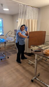 A woman in medical scrubs smiles and waves while wearing blue gloves in a hospital room, standing beside hospital beds, a wooden overbed table, and other medical furniture.