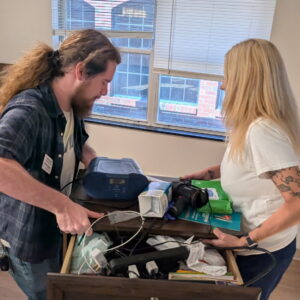 Two people stand beside an open drawer filled with various items, including cables and medical supplies, in a room with large windows and wooden floors. Both are looking into the drawer and organizing its contents.
