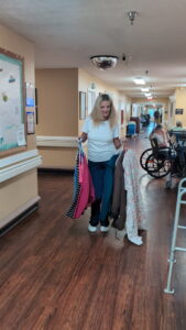 A woman stands in a hallway holding several coats; behind her, elderly people sit in wheelchairs. The setting looks like a care facility, with a bulletin board on the wall and a walker nearby.