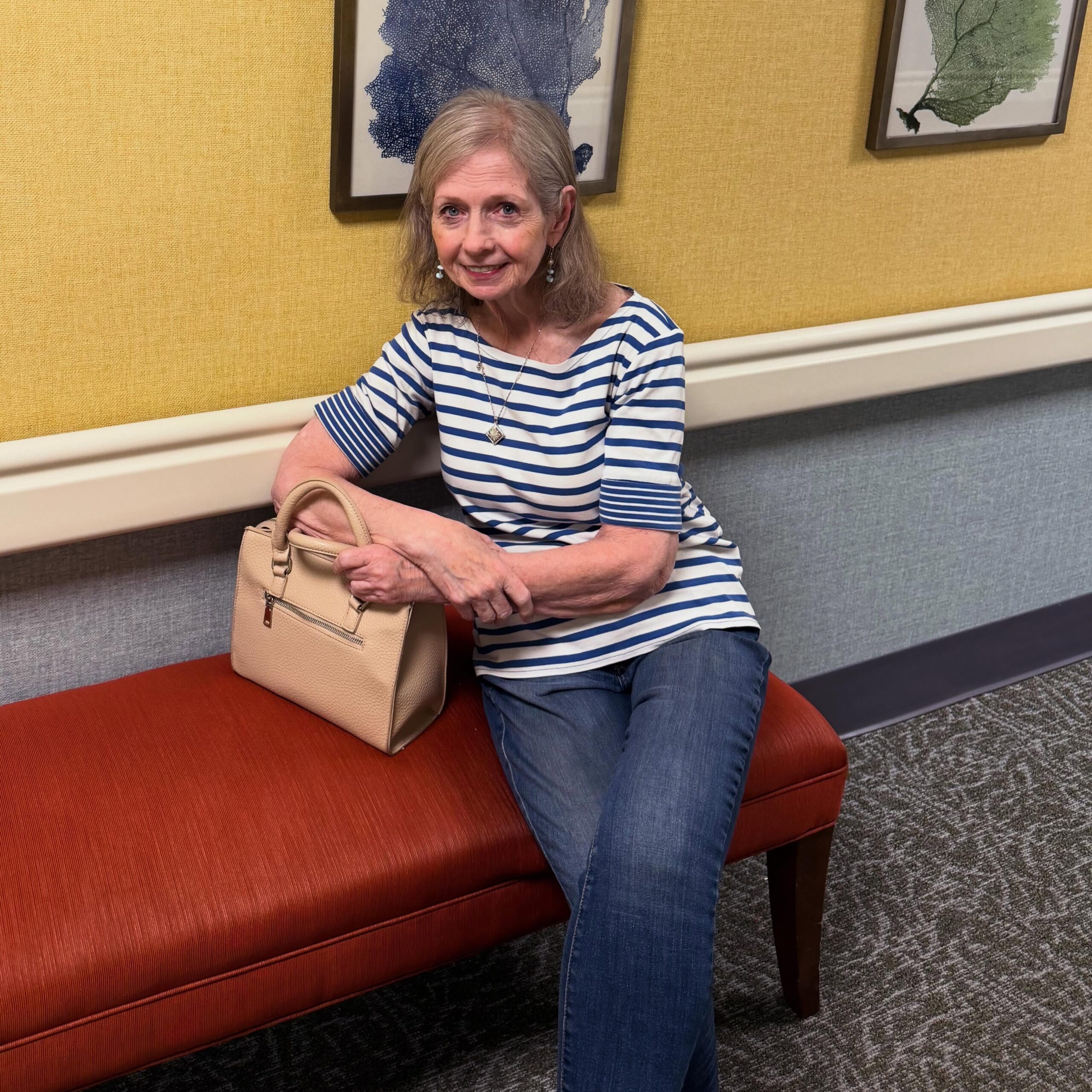 An older woman with shoulder-length gray hair, wearing a striped shirt and jeans, sits on an orange bench with a beige handbag in her lap at Wesley Manor memory care. The room features yellow and gray walls adorned with framed artwork.
