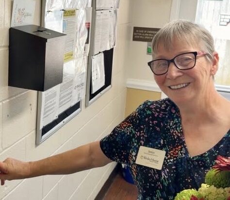 Smiling woman wearing glasses and a floral top clocks in using a fingerprint scanner on a wall. She holds a bouquet of red flowers and stands in a brightly lit room with bulletin boards in the background.