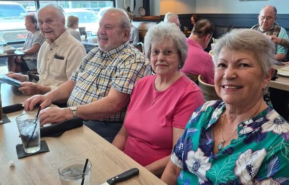 Four older adults, residents of Wesley Manor Independent Living, sit together at a restaurant table, smiling at the camera. Two women are in front, with two men behind them. Drinks and utensils are on the table, capturing a joyful moment.