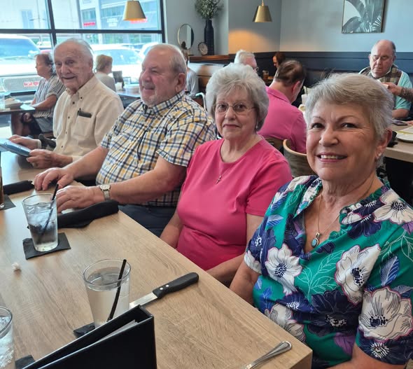 Four older adults, residents of Wesley Manor Independent Living, sit together at a restaurant table, smiling at the camera. Two women are in front, with two men behind them. Drinks and utensils are on the table, capturing a joyful moment.