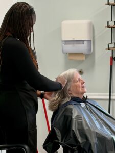 In a modern salon setting at Assisted Living at Wesley Manor, a hairstylist with long braids styles the hair of a seated older woman with gray hair, who is wearing a black cape and appears relaxed.