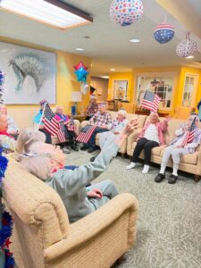In a cozy Wesley Manor memory care room adorned with patriotic balloons and flags, a group of smiling seniors wave American flags, reaching out joyfully as they celebrate together.