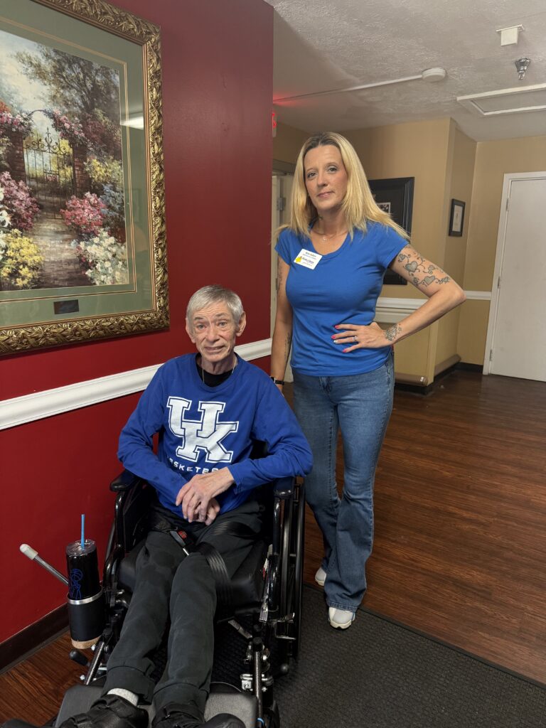 An older man in a wheelchair wearing a Kentucky shirt sits next to a woman in a blue shirt and jeans standing with her hand on her hip in a hallway with a framed painting on the wall.