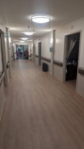 A long, well-lit hallway with wood flooring, doors on both sides, and an open room on the right. There are people and cleaning carts visible at the far end of the hall.
