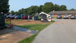 A small construction site with a Bobcat loader, a green dumpster, two people standing nearby, several parked cars, and a brick building in the background surrounded by trees.