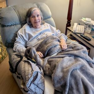 An older woman with gray hair sits in a recliner, covered with a gray blanket. She smiles at the camera. A phone, remote, water glass, and other items are on a table beside her.
