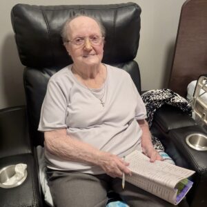 An elderly woman with glasses sits in a recliner, holding a pencil and a word search book, smiling at the camera. A tissue box sits beside her.