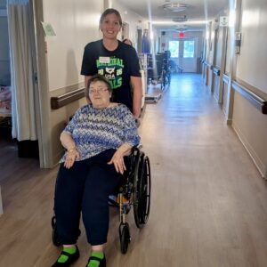 A younger woman stands behind an older woman in a wheelchair, smiling in a well-lit hallway of what appears to be a care facility.