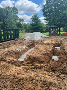 A construction site with dug trenches filled with gravel, a large pile of gravel, a green dumpster, trees, and a gazebo with people in the background under a partly cloudy sky.