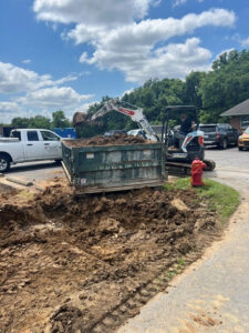 A small excavator loads dirt into a dumpster next to a white pickup truck. A large hole is in the foreground, with a red fire hydrant nearby. Trees and parked cars are in the background under a partly cloudy sky.